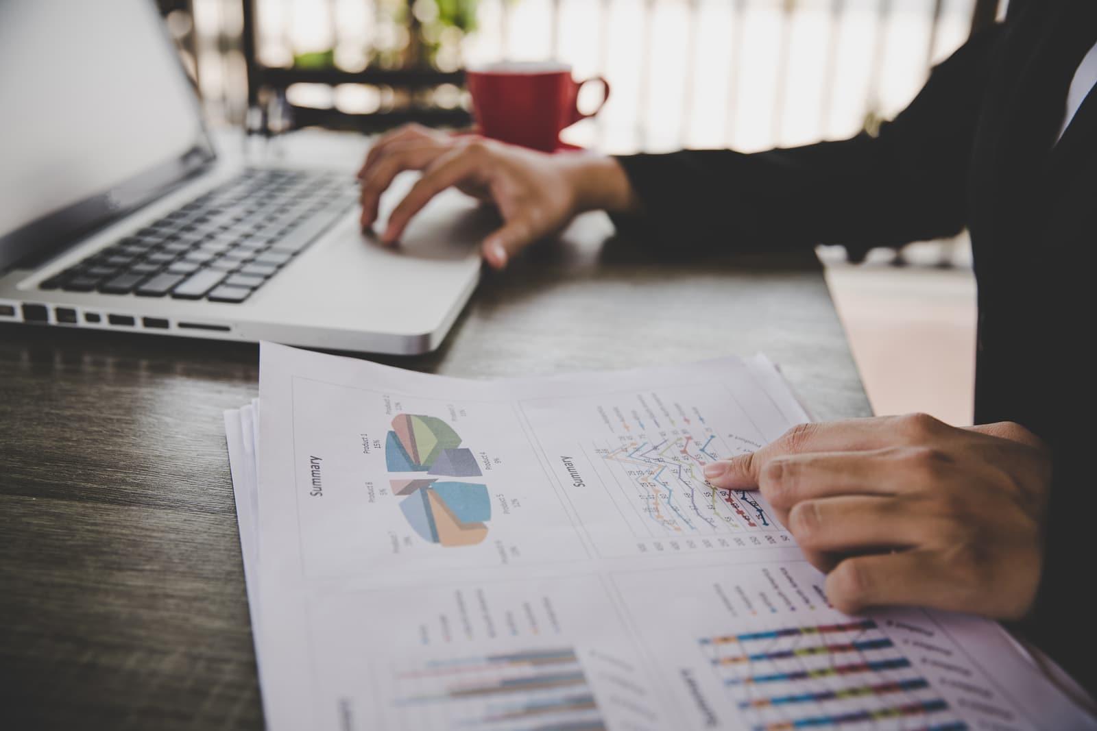 Person working at a laptop while reviewing printed charts and financial summaries on a desk